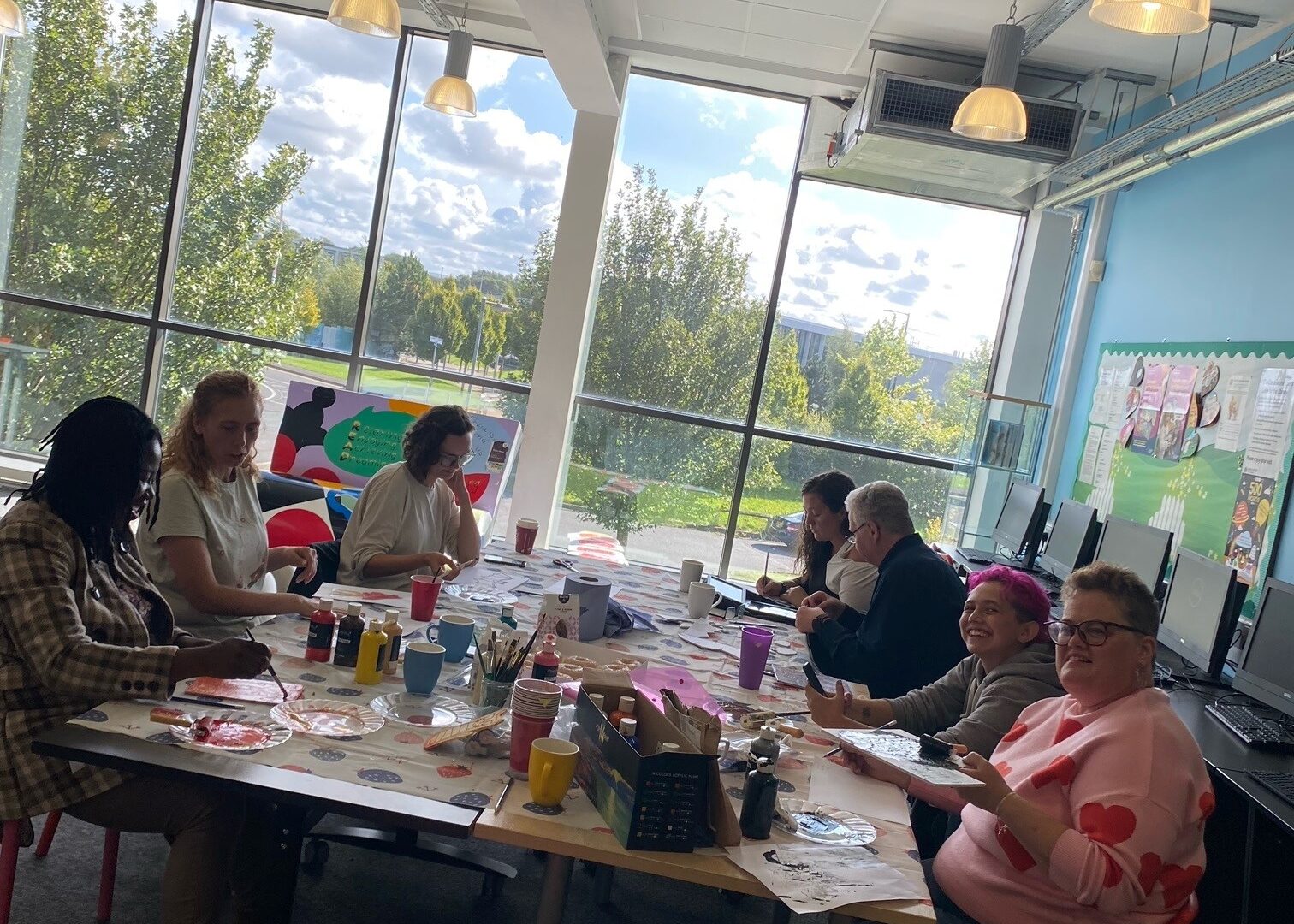 A group of 7 sit around a large table full of craft materials, including paint, large window overlooking trees behind them.